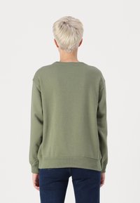 Olivově zelená mikina s volným střihem, žebrovanými manžetami a lemem. Měkká látka s hladkým povrchem. Nosená s modrými džíny.