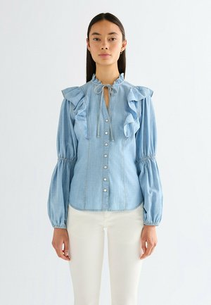 Lyseblå denimbluse med flæsekant ved kraven og ærmerne, med knaplukning foran, bindebåndslukning og opsamlede manchetter.