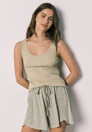 Giovane donna con capelli castani che indossa una canotta beige e pantaloncini con coulisse e motivo, in piedi contro una parete chiara e semplice.