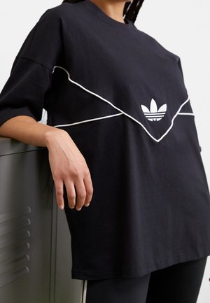 Henkilö pukeutuneena ylisuurikokoiseen mustaan Adidas-paitaan, jossa valkoiset koristeet ja logo, nojaa harmaaseen lukittavaan kaappiin, kuvattu kaulan ja reiden kohdalta.