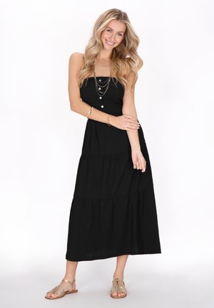 Schwarzes, schichtweise gestaltetes Maxikleid mit einem taillierten Oberteil mit Knöpfen, einem strukturierten Stoff und einer fließenden Silhouette. Kombiniert mit Riemchensandalen.