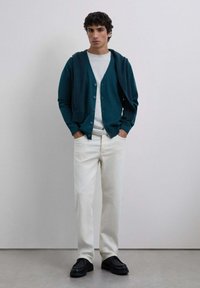 Cardigan teal cu nasturi, purtat peste un tricou gri, asortat cu pantaloni albi și pantofi negri, stând pe un fundal neutru.