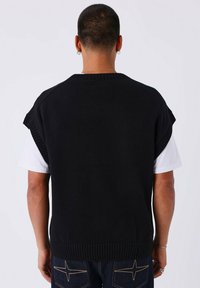 Jersey negro de punto sin mangas con bordes acanalados, cuello redondo y diseño de hombros caídos, llevado sobre una camiseta blanca.