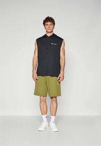 Svart ermeløs hettegenser med logo, kombinert med olivengrønne shorts og hvite joggesko; har snor og lommer.