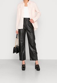 Mujer vestida con pantalones de cuero negros, loafers de plataforma negros, blusa blanca, camisa rosa claro y sosteniendo un bolso negro con cadenas doradas.