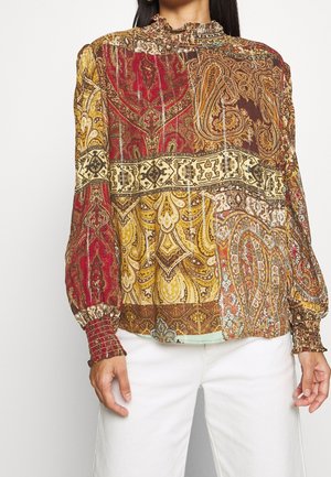 Kvinde iført langærmet patchwork-bluse med indviklede røde, gyldne og brune paisley- og geometriske mønstre kombineret med hvide bukser.