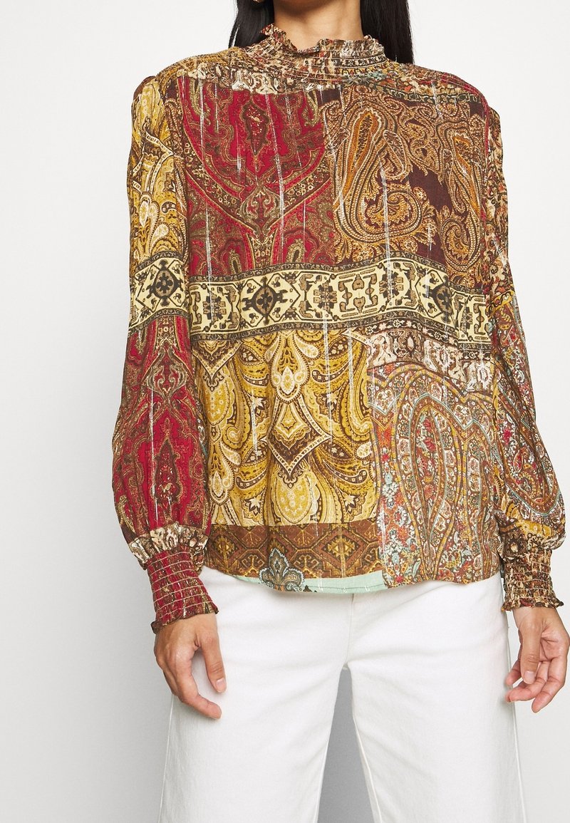 Femme portant un chemisier patchwork à manches longues avec des motifs complexes paisley rouges, dorés et bruns, associé à un pantalon blanc.