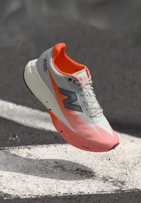 Chaussure de course New Balance en gris, avec des accents orange, une tige en mesh légère, une semelle en mousse rembourrée et un logo distinctif sur le côté.
