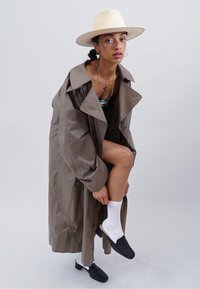 Langer, olivgrüner Trenchcoat mit breitem Kragen, getragen über einem schwarzen Outfit. Schwarze Slipper, weiße Socken und ein Strohhut.