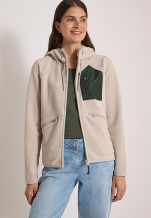 Kvinde iført en beige hættetrøje med lynlås og en grøn brystlomme, grøn skjorte og lyseblå jeans, stående foran en ensfarvet baggrund.