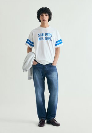 Joven de pie con camiseta blanca con texto "Scalpers Ath. Dept.", jeans azules, zapatos oscuros, sosteniendo una chaqueta gris sobre el brazo.