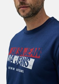 Námořnická modrá tričko s kulatým výstřihem, které obsahuje silný červený a bílý grafický text: "SALSA JEANS DENIM ARTISANS." Hladká bavlněná textura.