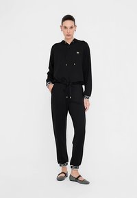 Juodas su gobtuvu sportinis kostiumas su raišteliais juosmenyje, kontrastingais rankogaliais su logotipo raštu ir paprasto dizaino lygiais šlepetėmis.