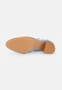 Suela de zapato azul de ante con una parte inferior de goma texturizada en tono marrón y un patrón geométrico. Presenta un tacón cuadrado y borde curvado. Logo de la marca visible.