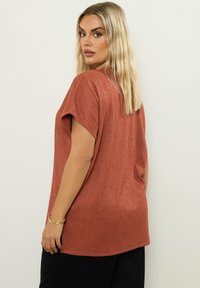 Lühikeste varrukatega roostes värvi topp, millel on tekstuuriga, sooniline kangas. Kõikuv lõige külgsugete ja mustade pükstega, koos lihtsa kuldse käevõruga.