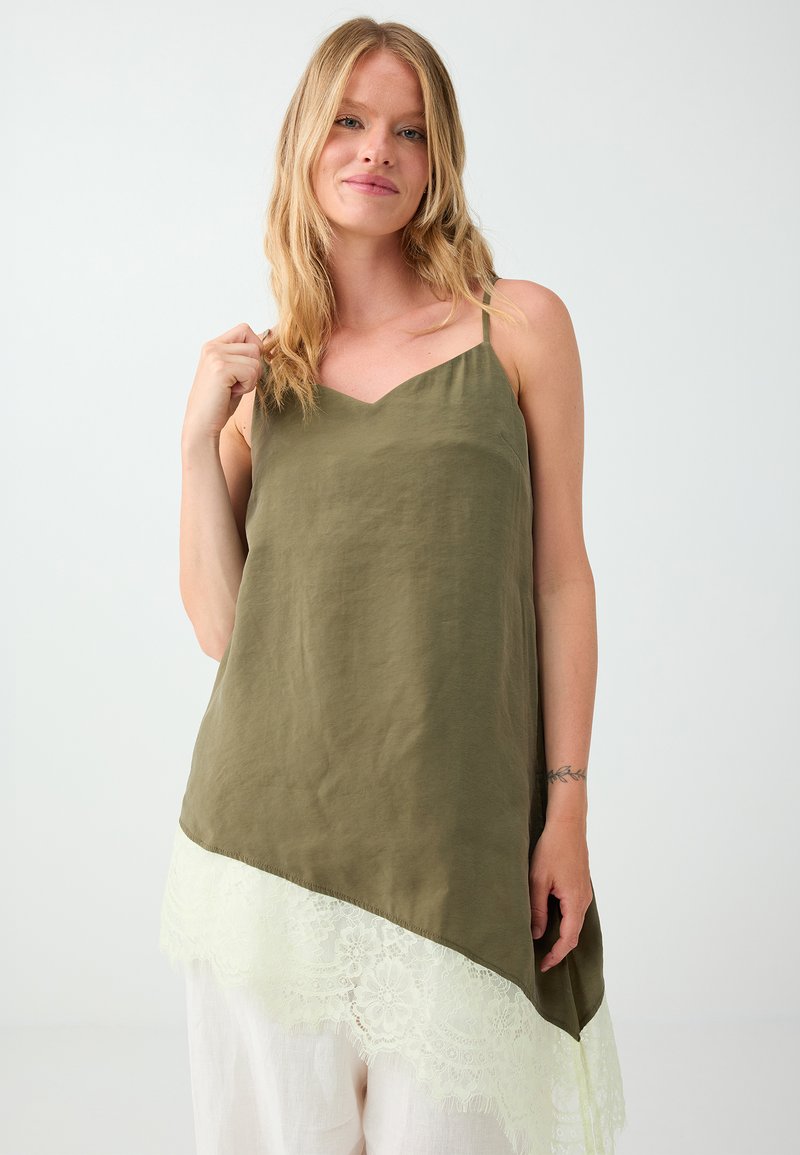 Femme aux cheveux blonds portant un haut sans manches vert olive avec un ourlet en dentelle blanche et un pantalon blanc sur fond clair uni.