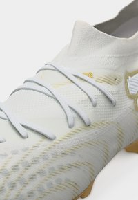 Baltas futbolo batelis su auksiniais akcentais, tekstūruotas megztas viršus, balti raišteliai ir formuoti smeigti ant šviesiai rudos pado.