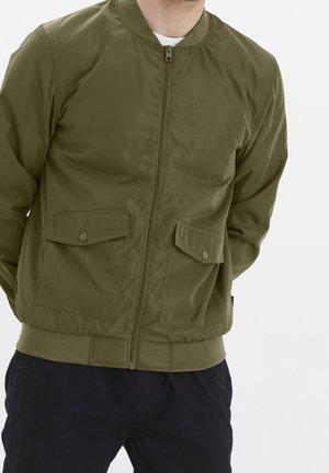 Homme portant une veste bomber vert olive zippée, avec deux poches à rabat boutonnées et un pantalon foncé sur un fond clair uni.