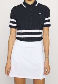 Svart ribbad polo med vita ränder, knäppbar krage, kombinerad med en vit kjol. Har en liten logotyp på skjortan.