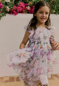 Witte jurk met kleurrijke vlinderafdrukken, gelaagd stof, ruches aan de mouwen en een verzamelde taille. Achtergrond met roze bloemen en groen.