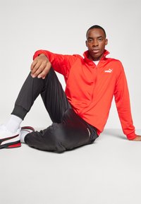 Rode zip-up jas met hoge kraag, zwarte sportbroek en witte sneakers. Model zit, waarbij het logo van de jas en de textuur van de broek zichtbaar zijn.
