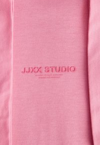 Tecido rosa claro com uma textura suave apresentando o logótipo em relevo "JJXX STUDIO". A costura é visível nas bordas.