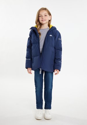 Mørkeblå puffjakke med gul hættefor, lynlås fortil og elastiske manchetter. Båret over en grå skjorte og parret med blå jeans og hvide sneakers.