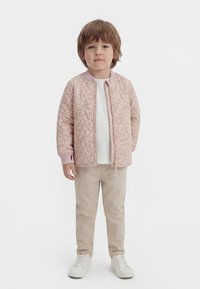 Majhen otrok stoji, oblečen v svetlo roza prešito jakno, belo majico, bež hlače in bele superge, na enobarvnem belem ozadju.