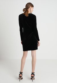 Rochie din catifea neagră cu mâneci lungi și o siluetă ajustată, având un guler rotund. Purtată cu sandale cu tocuri negre și curelușe.