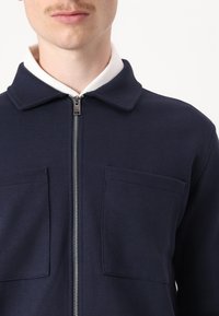 Navy zip-up jacket with a textured fabric, two frontal pockets, and a white collar, featuring a metal zipper detail.