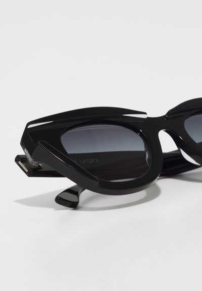Black sunglasses with angular, geometric design, featuring gradient lenses. Glossy plastic frame with distinctive side accents.