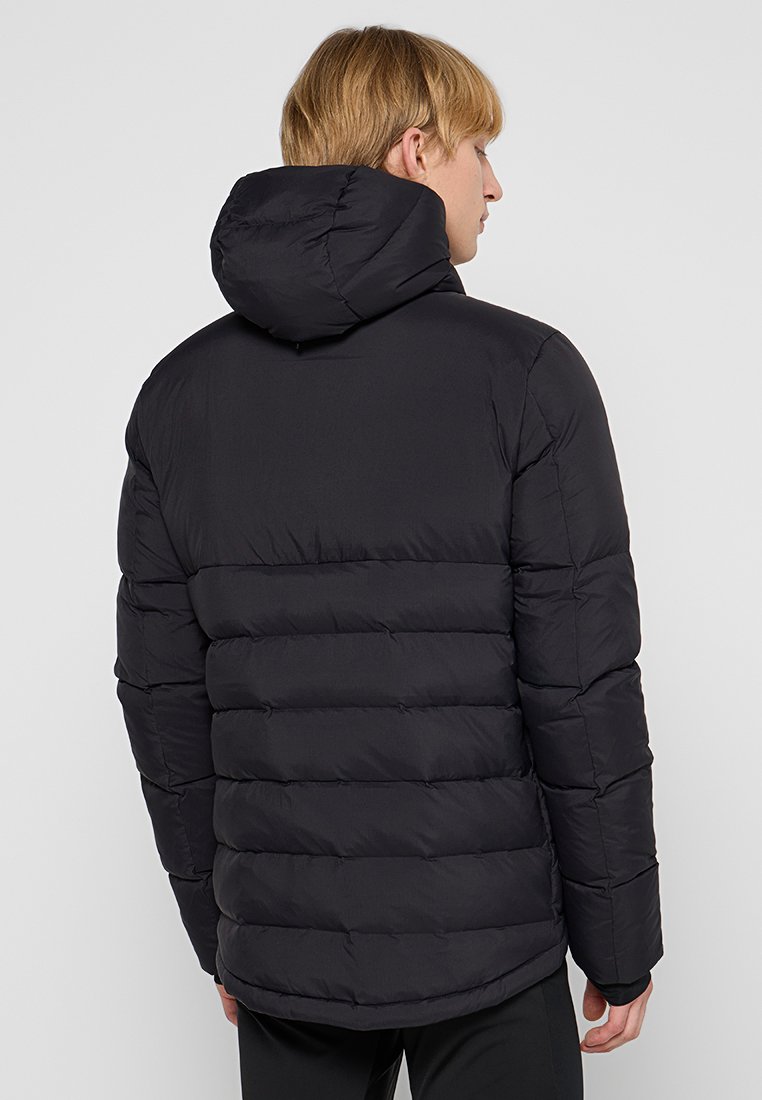 Ein blonder Mann in einer schwarzen gepolsterten Kapuzenjacke, der mit dem Rücken zu einer schlichten hellen Hintergrund steht.