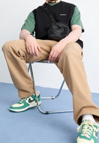 Gilet nero sopra una camicia verde, pantaloni cargo color sabbia e sneakers Nike verde e bianco. Piccola borsa a tracolla nera; seduto su una sedia cromata.