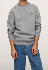 Sudadera gris de cuello redondo hecha de una mezcla de algodón, con puños y dobladillo acanalados. Pequeño logotipo circular en el lado izquierdo del pecho. Llevada con vaqueros azules.