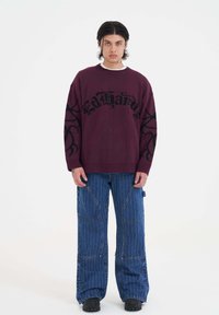 Vínově červený oversized svetr s černým grafickým textem a motivy na rukávech, spárovaný s modrými pruhovanými širokými kalhotami a černými botami.