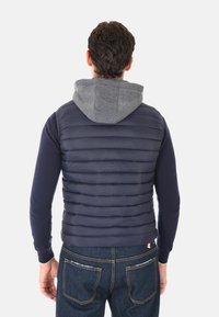 Uomo che indossa un gilet imbottito blu navy sopra un maglione blu navy con cappuccio grigio, in piedi di spalle contro uno sfondo bianco semplice.