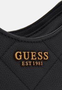 Czarna pikowana torebka wykonana z teksturowanego materiału z złotym logo, które brzmi "GUESS EST 1981." Posiada gładkie krawędzie i wyraźne przeszycia.