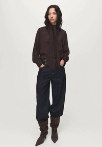 Blouse marron foncé avec un col noué, associée à un jean oversized en denim foncé. Le mannequin porte des bottes brunes, amples et montant jusqu'aux genoux. Fond simple.