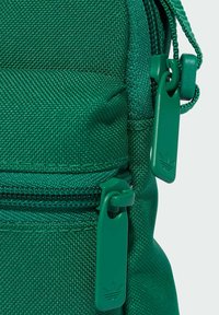 Zöld szövet táska, amely texturált anyagból készült, két cipzáras rekesszel és zöld cipzárhúzókkal rendelkezik, melyeken logó van dombornyomva.