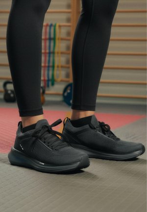 Osoba nosící černé sportovní boty a černé legíny stojí na podlaze posilovny, přičemž v pozadí jsou vidět kettlebells a posilovací pásy.