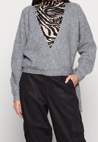 Grå stickad sveptröja med djup V-ringning, texturerad tyg och långa ärmar, lagerpå en svart och beige zebra-tryckt topp.