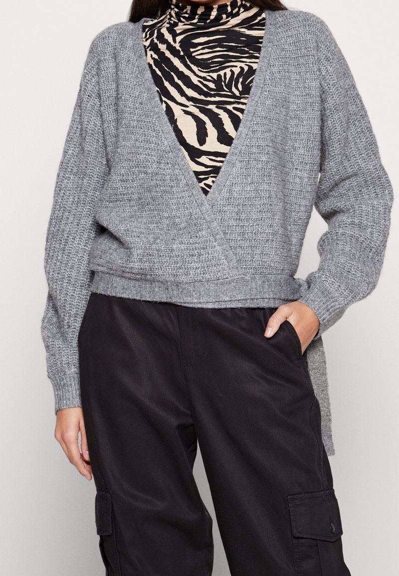 Grå stickad sveptröja med djup V-ringning, texturerad tyg och långa ärmar, lagerpå en svart och beige zebra-tryckt topp.