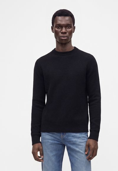 Man wearing a black long-sleeve sweater and blue jeans standing against a plain white background.