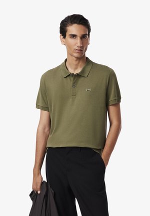 Oliva-grünes Poloshirt aus strukturiertem Stoff, mit einem Kragen, zwei Knöpfen und einem kleinen Logo auf der Brust. Kombiniert mit schwarzen Hosen.