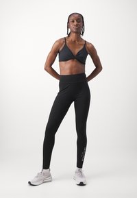 Black sports bra with a twisted front, paired with high-waisted black leggings. Model wears light sneakers for a complete athletic look.