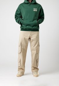 Groene katoenen hoodie met een voorzak en tekstlogo, gecombineerd met beige cargobroeken met meerdere zijzakken. Neutrale sneakers maken de look compleet.