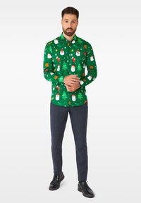 Camicia verde a maniche lunghe con un motivo natalizio giocoso che presenta Babbo Natale, alberi di Natale e decorazioni. Abbinata a pantaloni scuri a trama e scarpe nere.