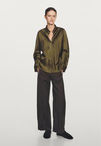 Satin, olive green button-up shirt with a relaxed fit and dropped shoulders, paired with dark brown wide-leg trousers and black shoes.