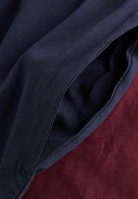 Granatowy materiał złożony na teksturowanym bordowym materiale, ukazujący szczegółowe przeszycia i miękkie fałdy tkaniny.