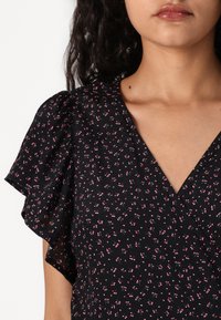 Blusa preta floral com mangas bufantes, apresentando um decote em V profundo e um padrão de pequenas flores cor-de-rosa com folhas verdes. Tecido macio e texturizado.